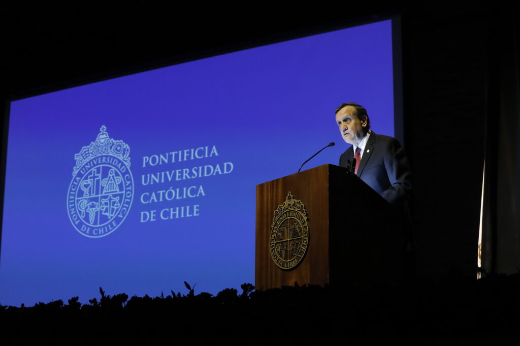 Fotografía del Rector Ignacio Sánchez en el podio con el escudo UC, de fondo se ve una pantalla azul con el logo de la universidad