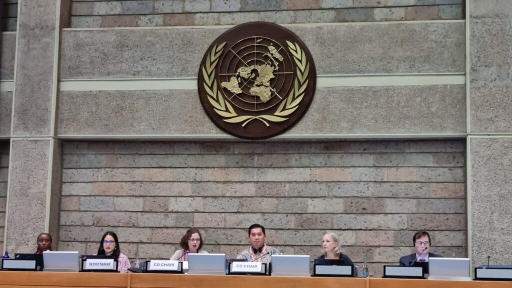 Fotografía de 6 personas sentadas en un gran mesón en altura, arriba se ve grande el logo de la Organización de las Naciones Unidas, ONU.