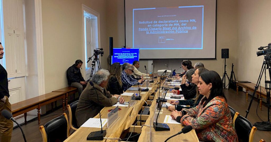 Fotografía de un grupo de personas sentadas con computadores en una mesa larga, de fondo se ve una presentación proyectada en un telón