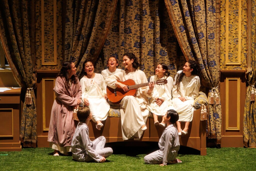 Fotografía de Vanesa Rojas sentada sonriendo con una guitarra en un escenario al rededor de un grupo de niñas y adolescentes