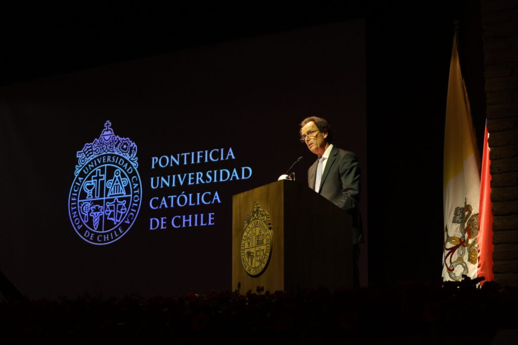 Fotografía del Rector UC Juan Carlos de la Llera en un escenario hablando frente al podio de madera con el escudo de la UC y de fondo una presentación con el logo oficial de la Pontificia Universidad Católica de Chile