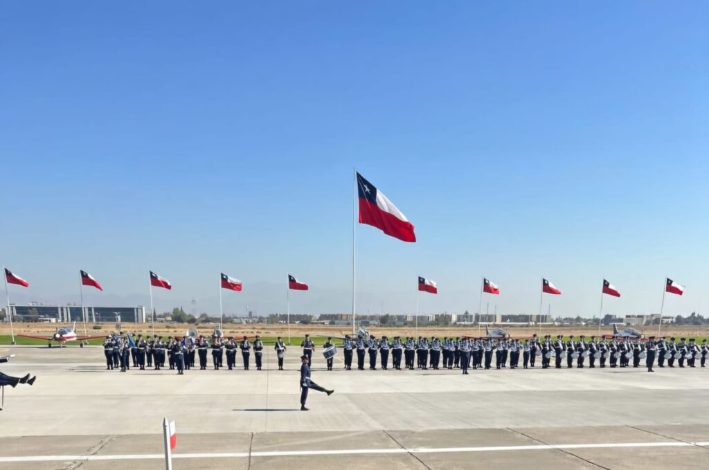 Fotografía donde se ve una persona desfilando frente a una fila de personas en uniforme, de fondo muchas banderas de chile pequeñas, y al centro una gran bandera del mismo país